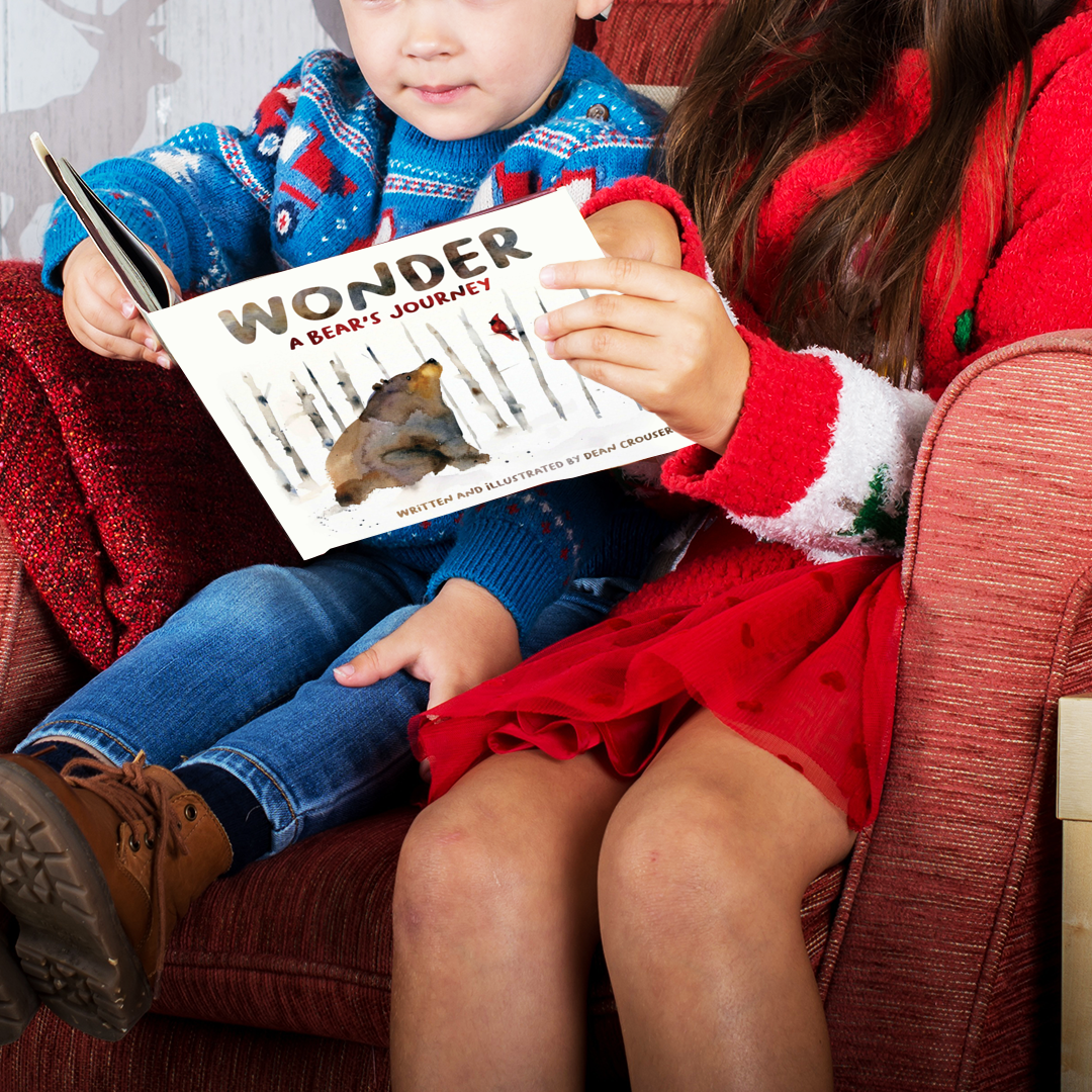 A young boy and an adult woman, seated in a chair together, reading the book “WONDER: A Bear’s Journey” together.